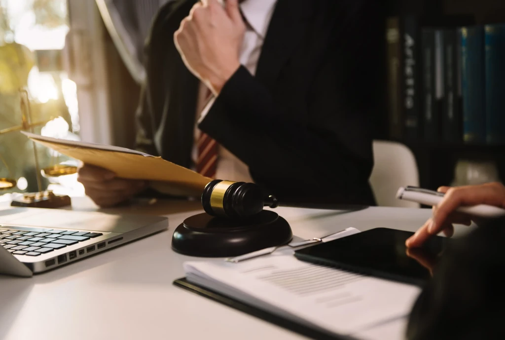 A lawyer at a desk holds documents, with a gavel and scales of justice nearby. The scene is serious, with a laptop and legal books in the background.