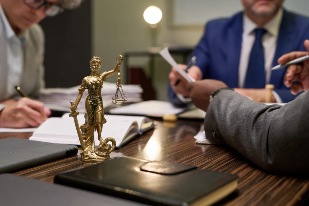 A small statue of Lady Justice is on a table surrounded by three people in suits, engaged in a serious meeting with documents and notebooks.