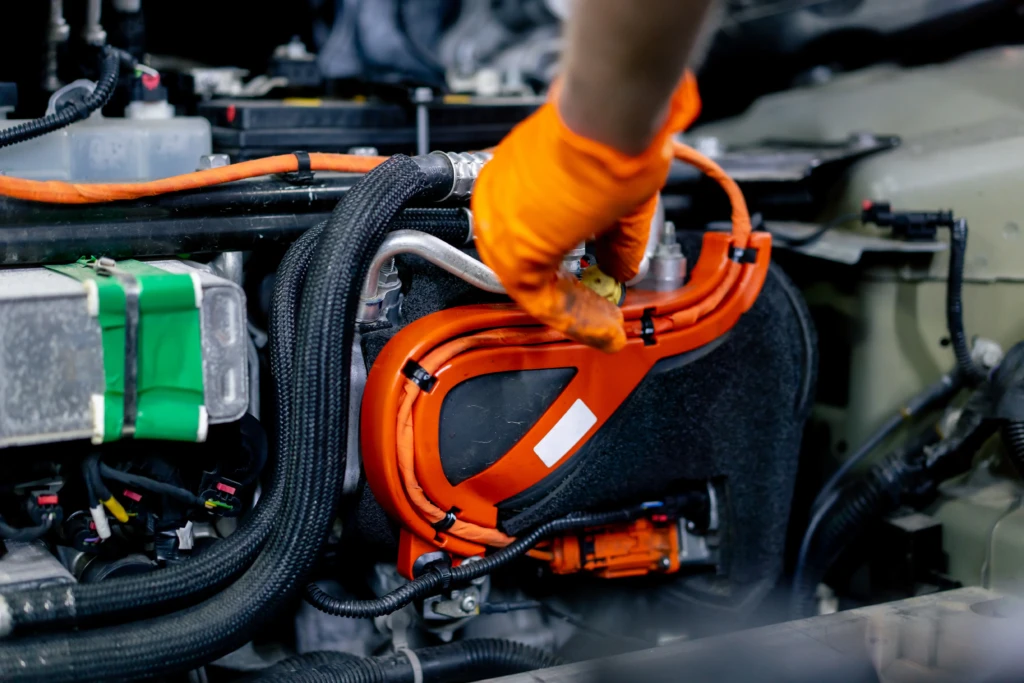 A person wearing orange gloves works on an electric car's engine, focusing on orange cables and components, conveying a technical and industrious tone.