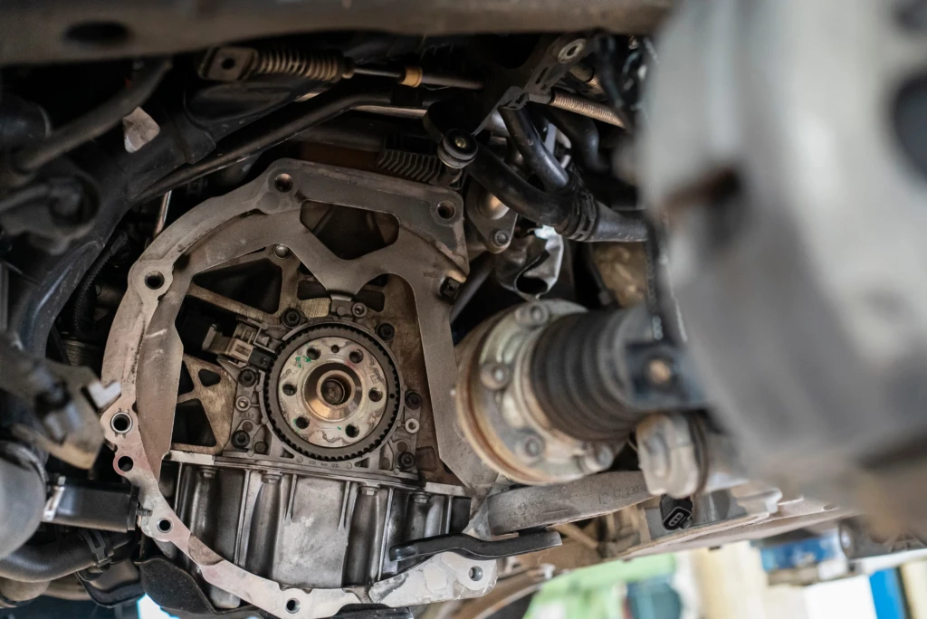 Close-up view of a car's transmission system from underneath, showcasing gears, drive shaft, and metal components in a complex mechanical assembly.