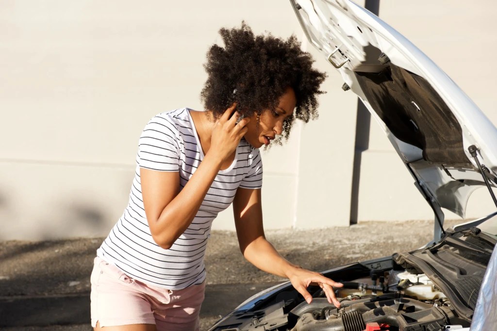 A woman stands beside an open car hood, examining the engine while talking on her phone. She appears focused and concerned, wearing a striped shirt and shorts.